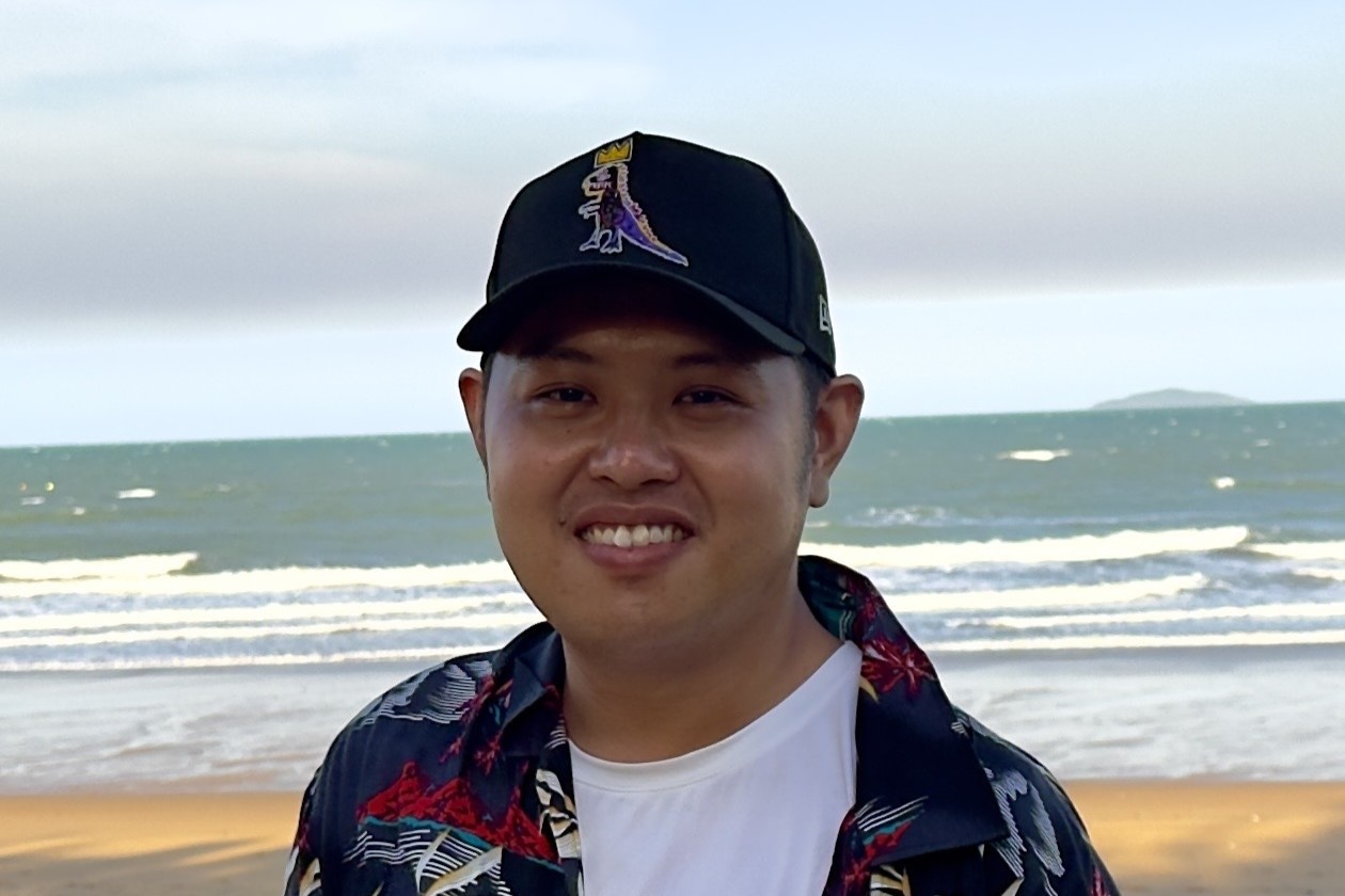 A close-up portrait of a smiling East Asian man wearing a black baseball cap with a dinosaur graphic and a patterned open shirt, standing in front of a beach with ocean waves.